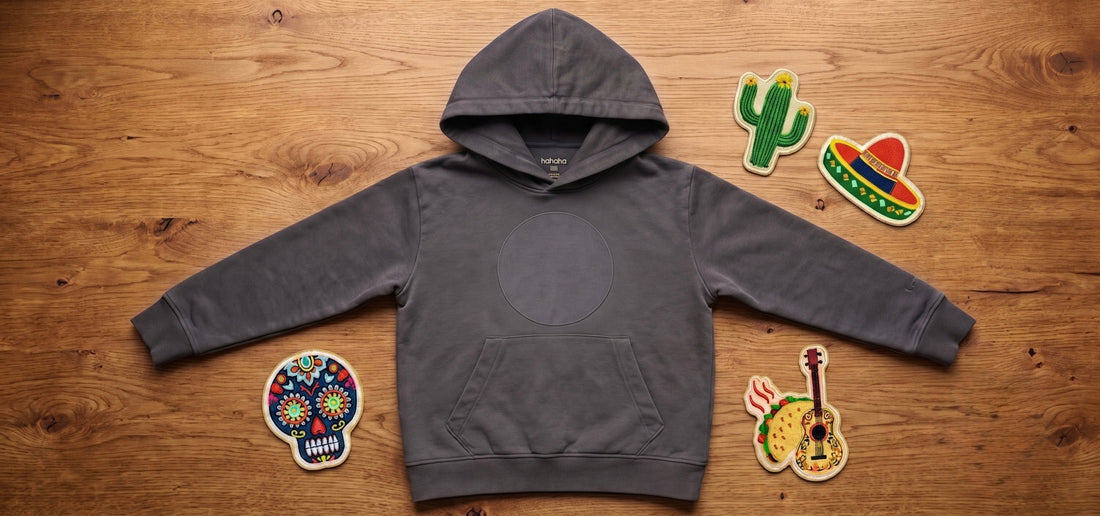 Black organic cotton kids hoodie with a patch area, surrounded by three swappable embroidered patches on a wood surface