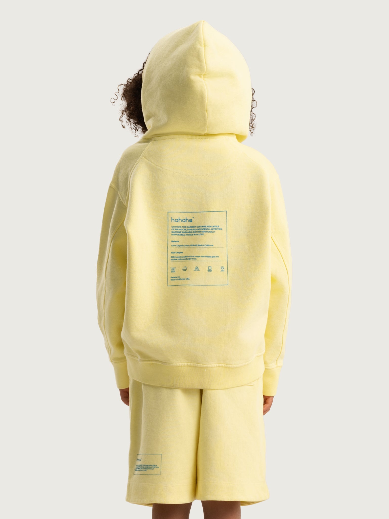 Back of light yellow Hahaha kids hoodie with CAUTION CONTAINS HIGH LEVELS OF SNUGGLES AND GIGGLES label